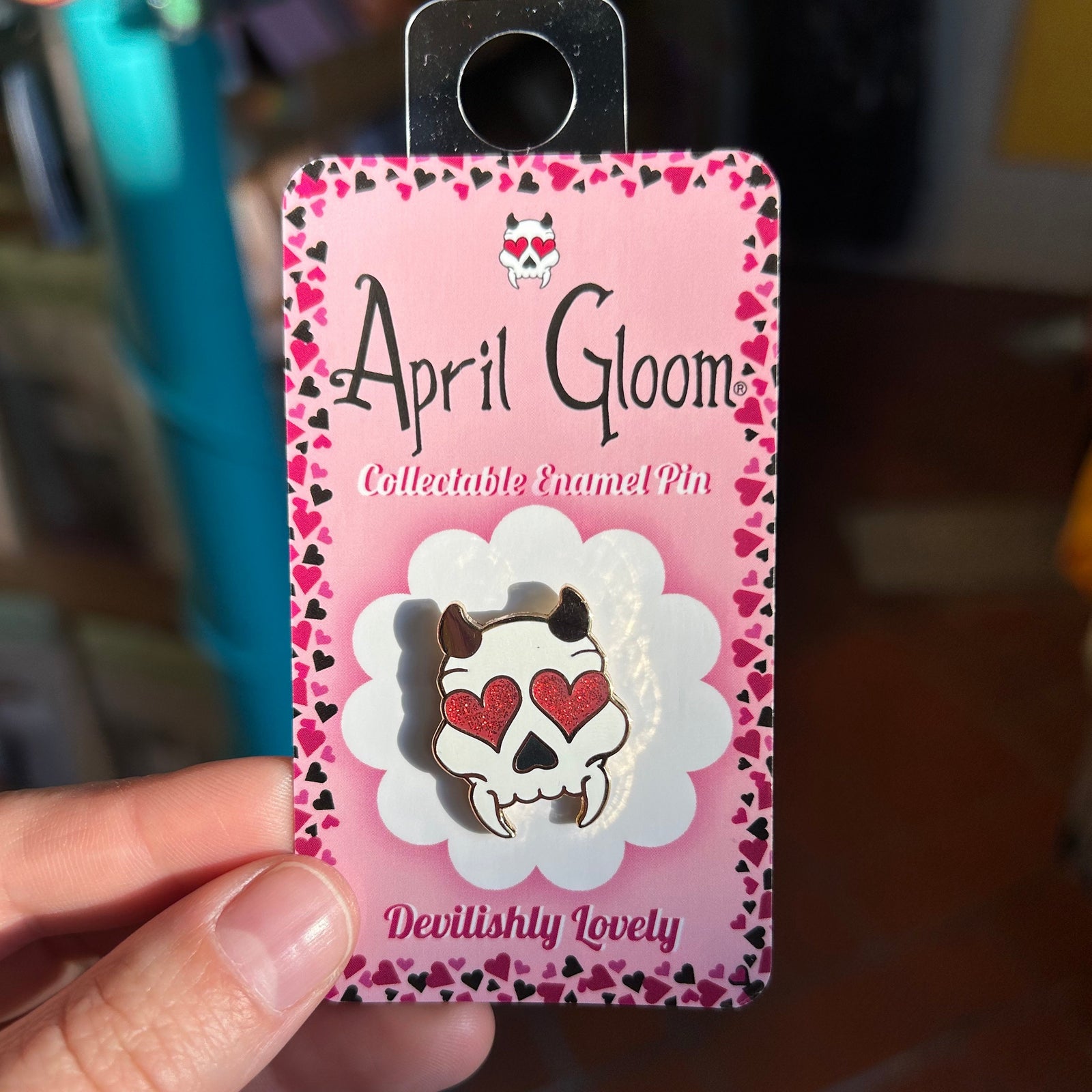 An enamel pin of a skull with heart eyes and horns on a pink backing card that says "April Gloom/Collectable Enamel Pin/Devilishly Lovely" held by a hand. 