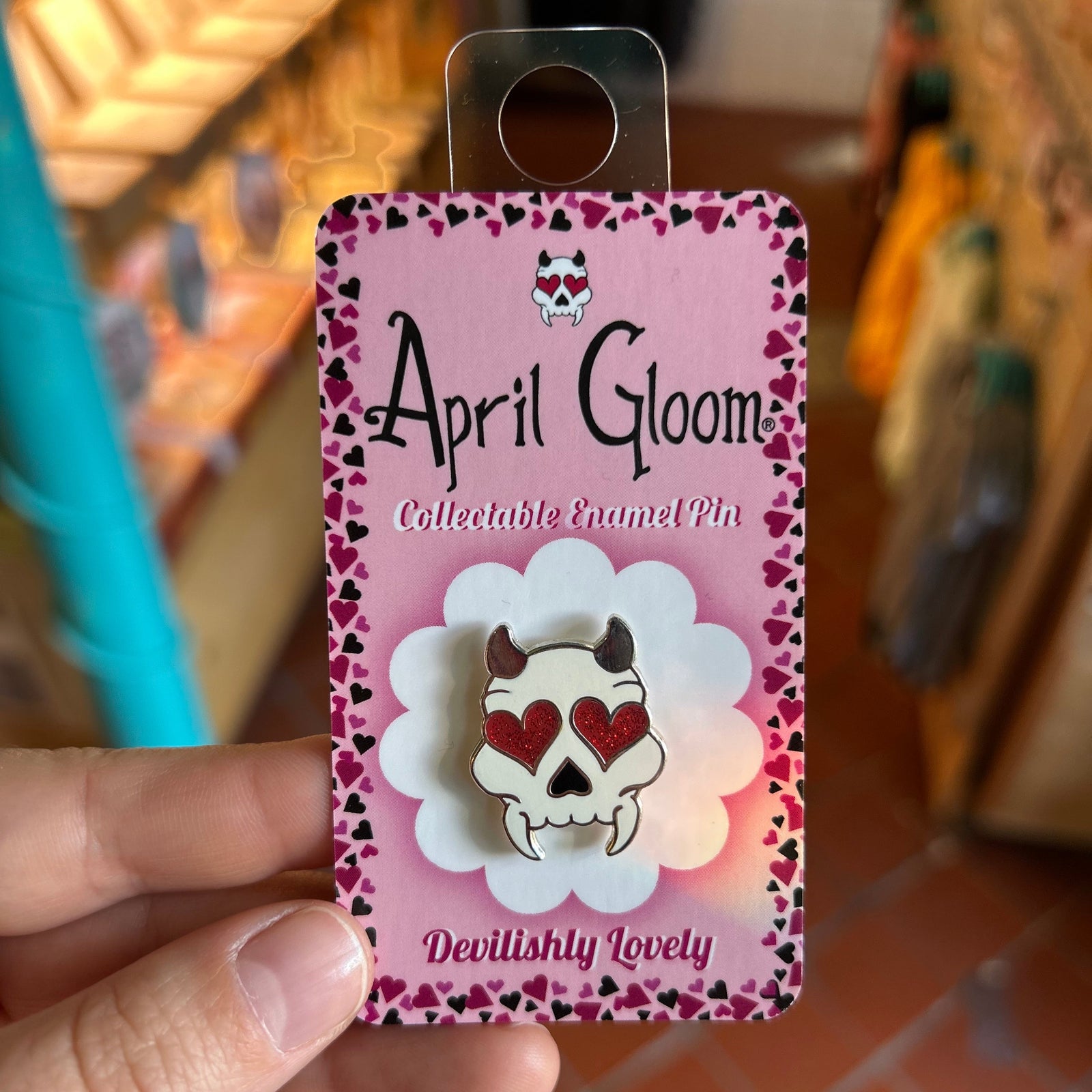 An enamel pin of a skull with horns and heart eyes on a pink backing card that says "April Gloom/Collectable Enamel Pin/Devlishly Lovely" held up by a hand