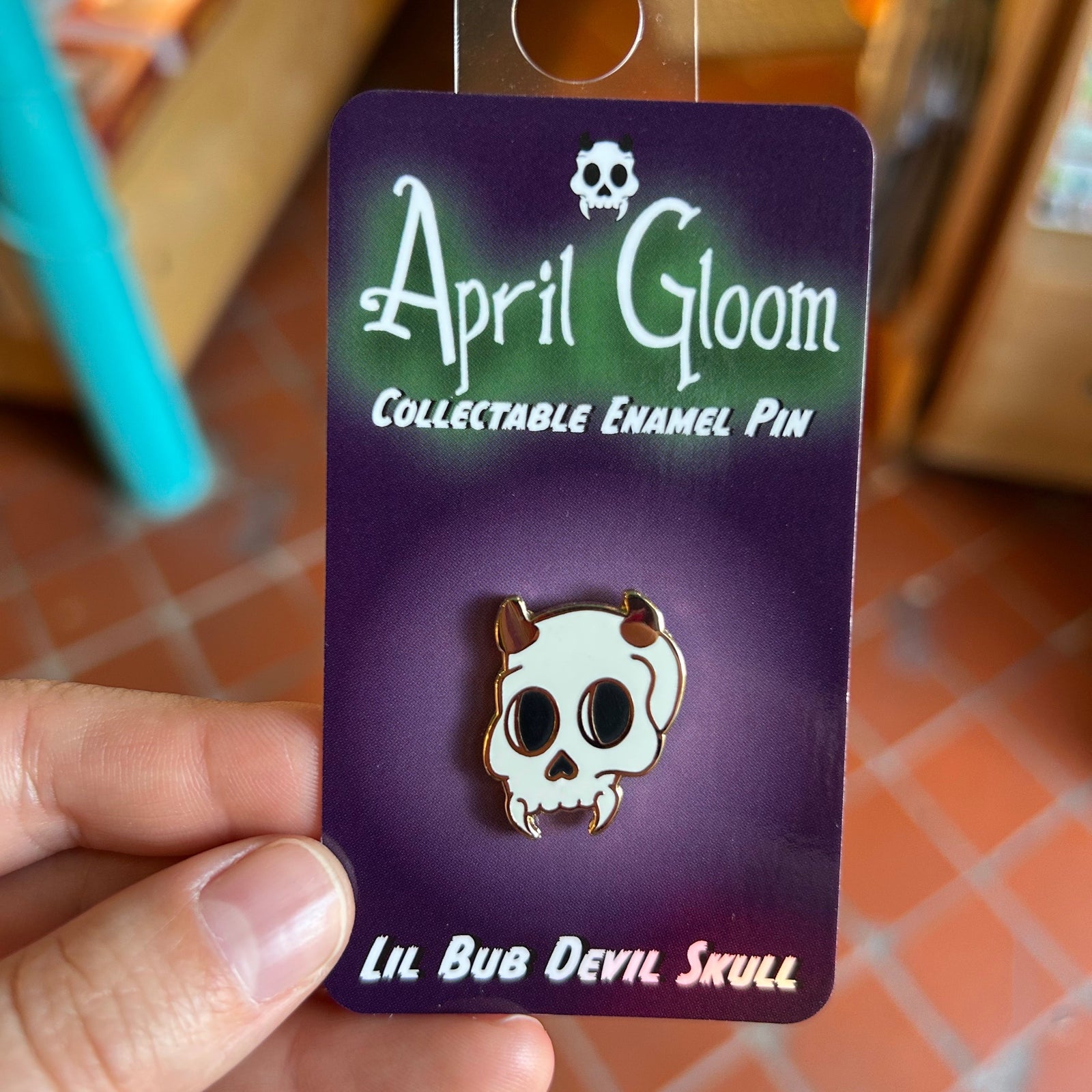 An enamel pin of a skull with horns on a purple backing card that says "April Gloom/Collectable Enamel Pin/Lil Bub Devil Skull" held up by a hand