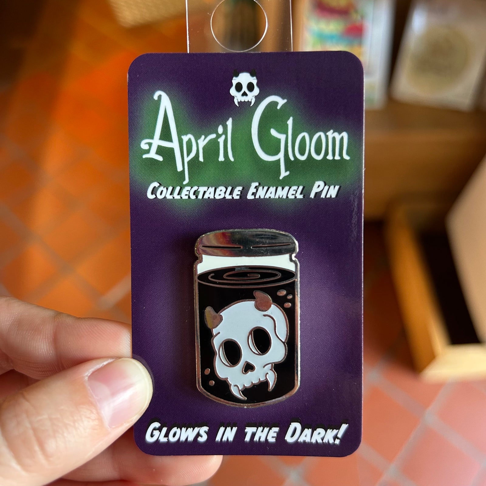 An enamel pin of a skull with horns inside a jar with black liquid on a purple backing card that says "April Gloom/Collectable Enamel Pin/Glows in the dark!"