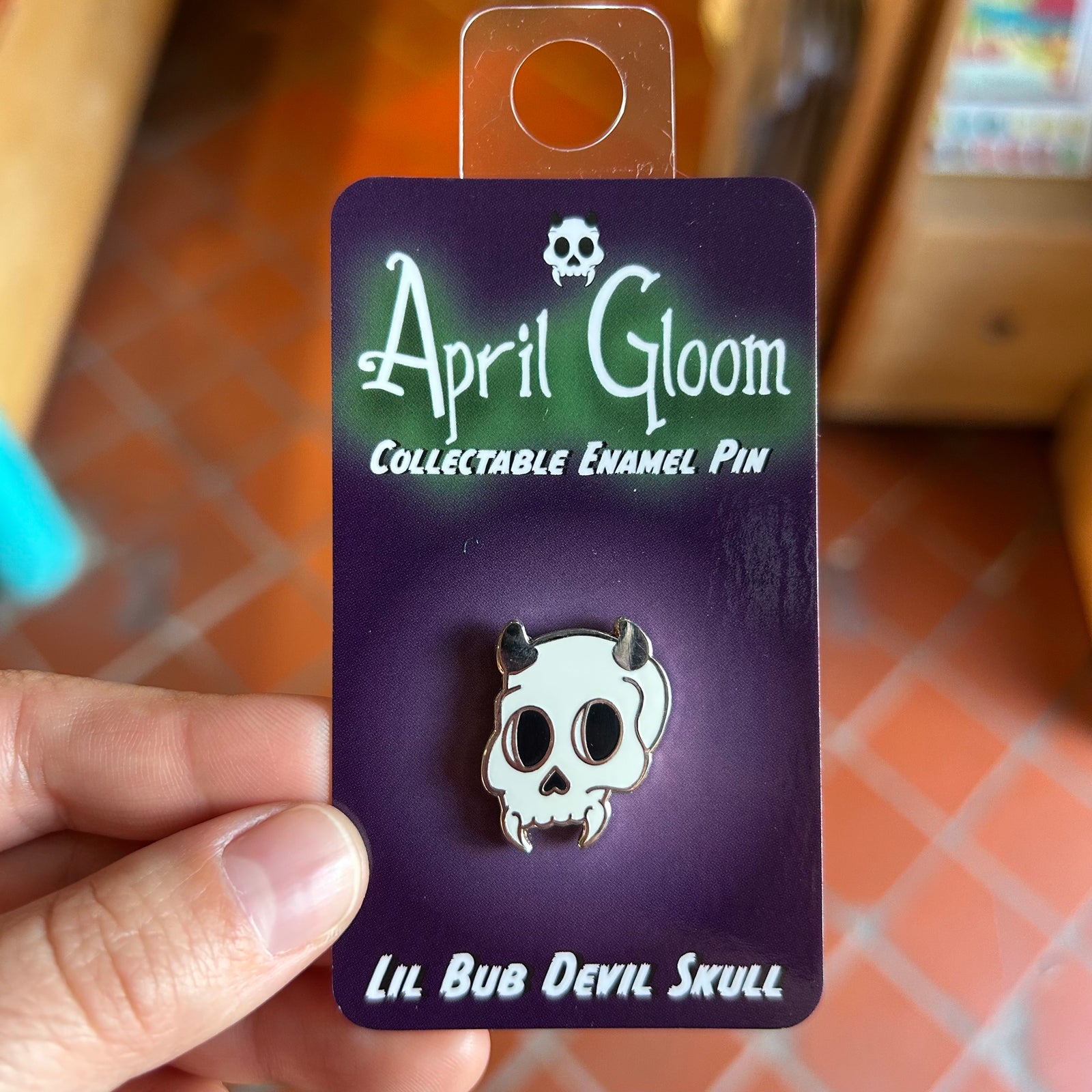 An enamel pin of a skull with horns on a purple backing card that says "April Gloom/Collectable Enamel Pin/Lil Bub Devil Skull" held up by a hand. 