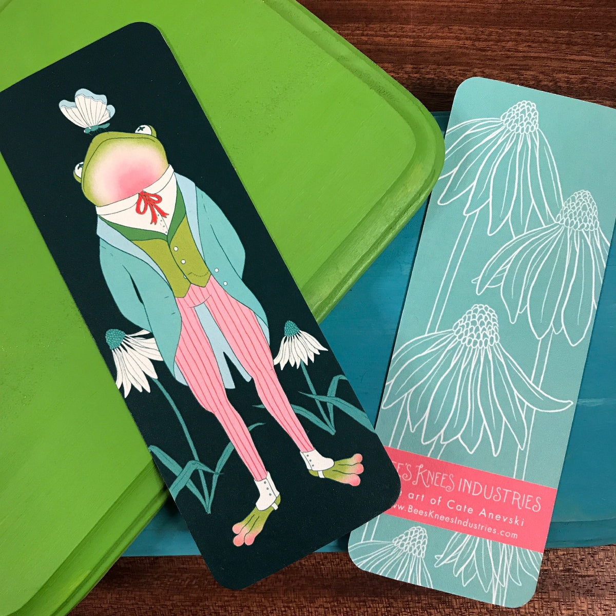 Bookmark that has a frog wearing a suit with a butterfly on his nose on one side and white daisy illustrations on a blue background on the other side.