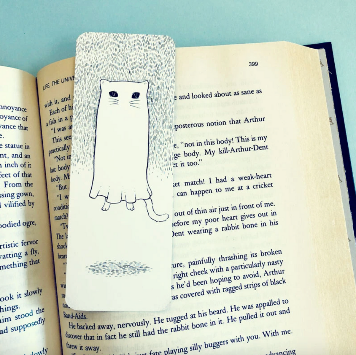A bookmark that has a black and white line drawing illustration of a cat-shaped ghost sitting on top of a page of a book. 