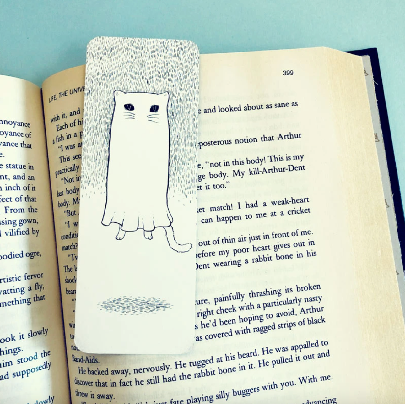 A bookmark that has a black and white line drawing illustration of a cat-shaped ghost sitting on top of a page of a book. 
