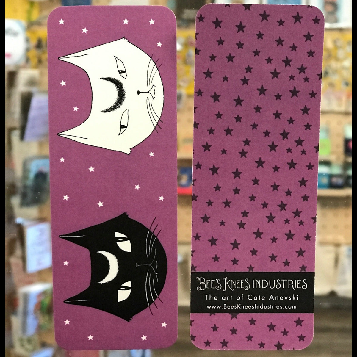 A bookmark that has two cats with crescent moon shapes on their foreheads on one side and black stars on a purple background on the other.