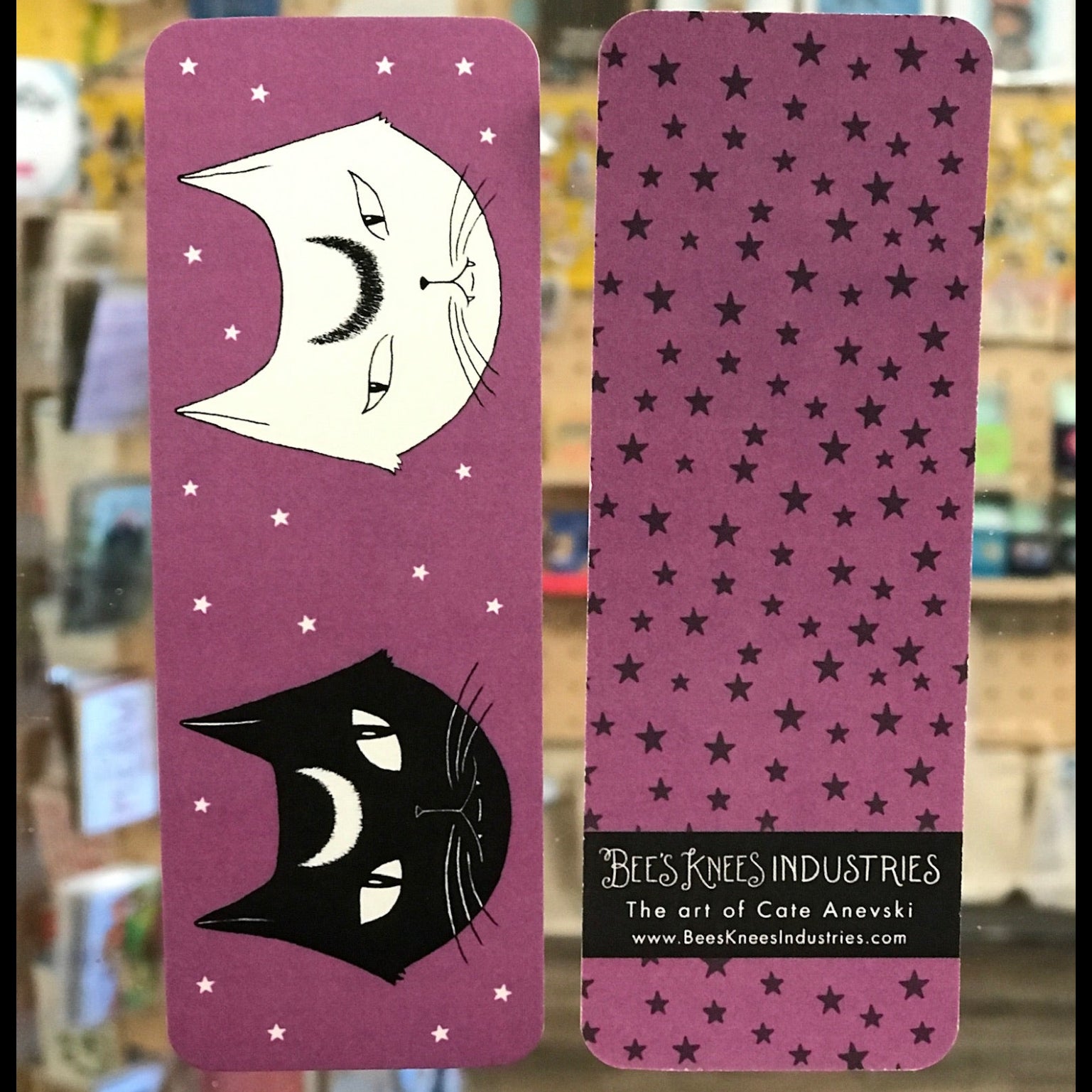 A bookmark that has two cats with crescent moon shapes on their foreheads on one side and black stars on a purple background on the other.