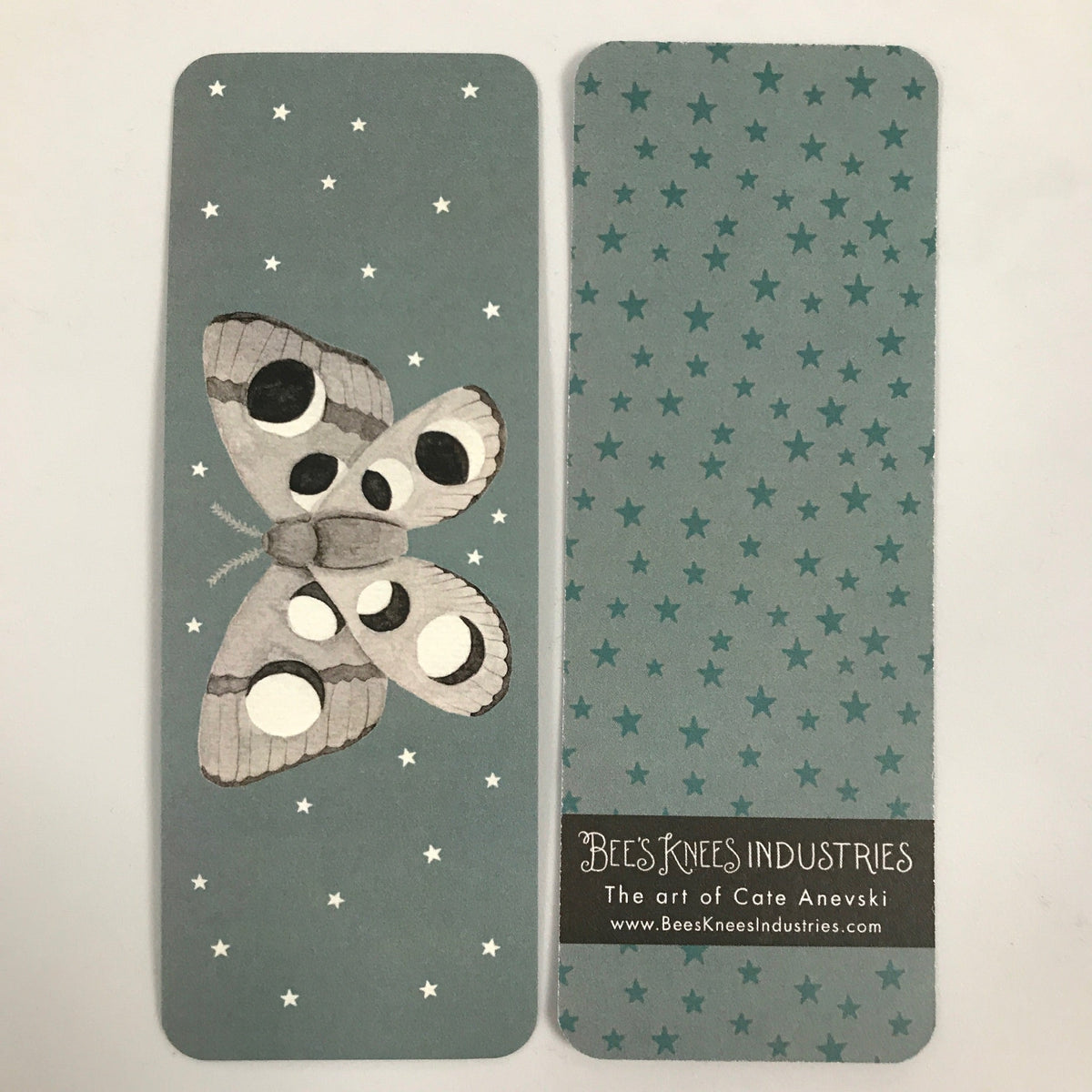 A bookmark with a moth with phases of the moon on its wings on one side and dark green stars on a light green background on the other. 