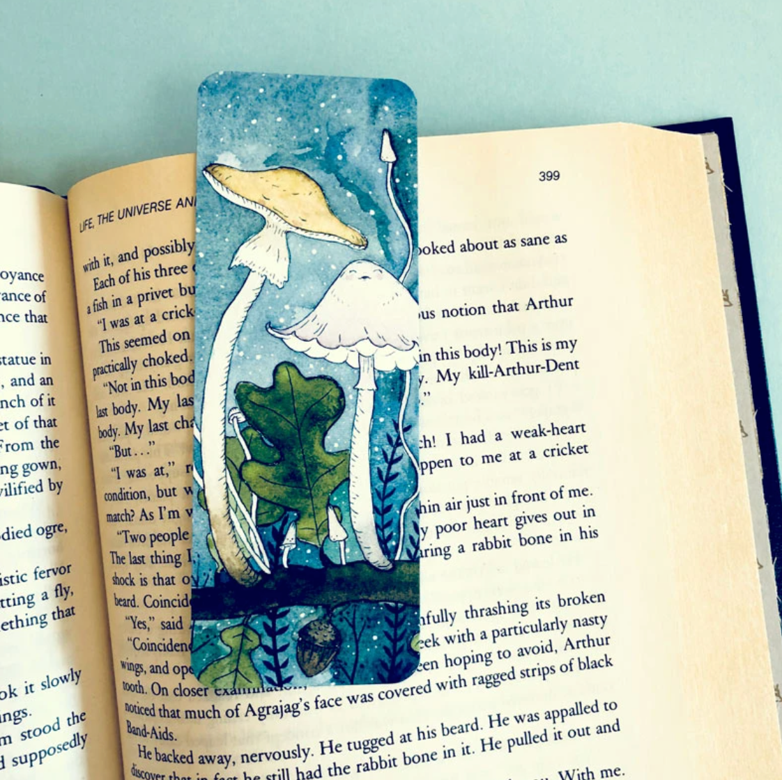 A bookmark with a watercolor illustration of two mushrooms on an oak tree branch against a starry sky. 