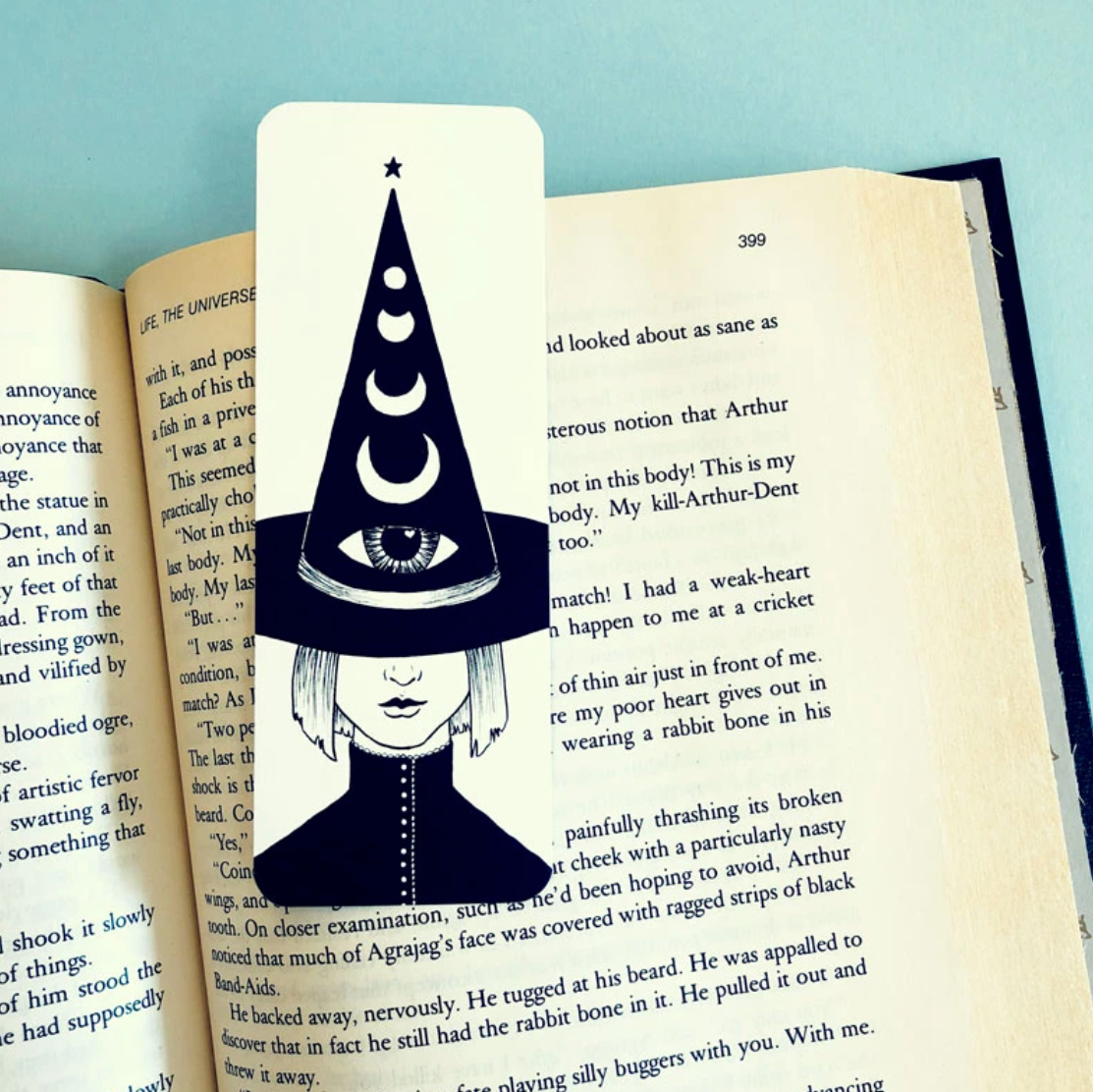 A bookmark that features an illustration of a witch wearing a hat that has a large eye and a series of crescent moons on it.