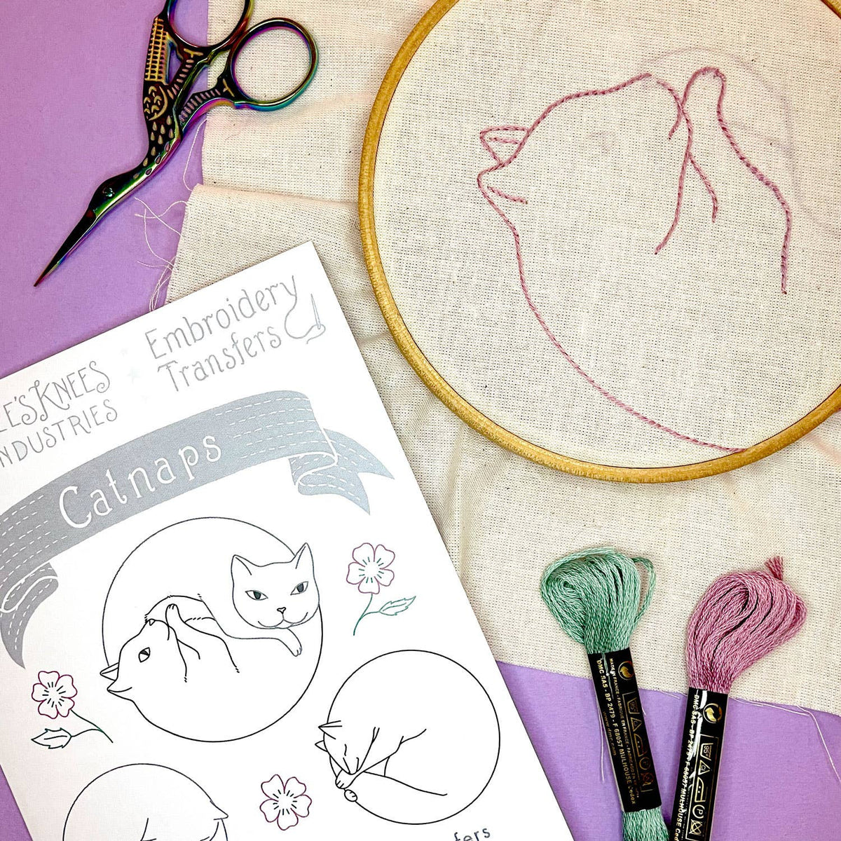 Embroidery hoop with cat design, scissors, and embroidery thread on a purple surface.