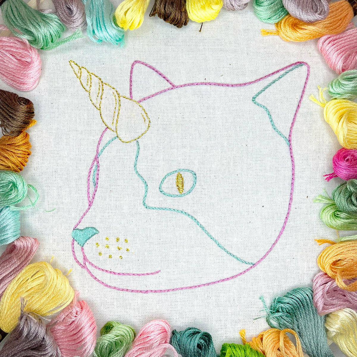 Embroidery hoop with a cat unicorn design surrounded by colorful floss skeins.