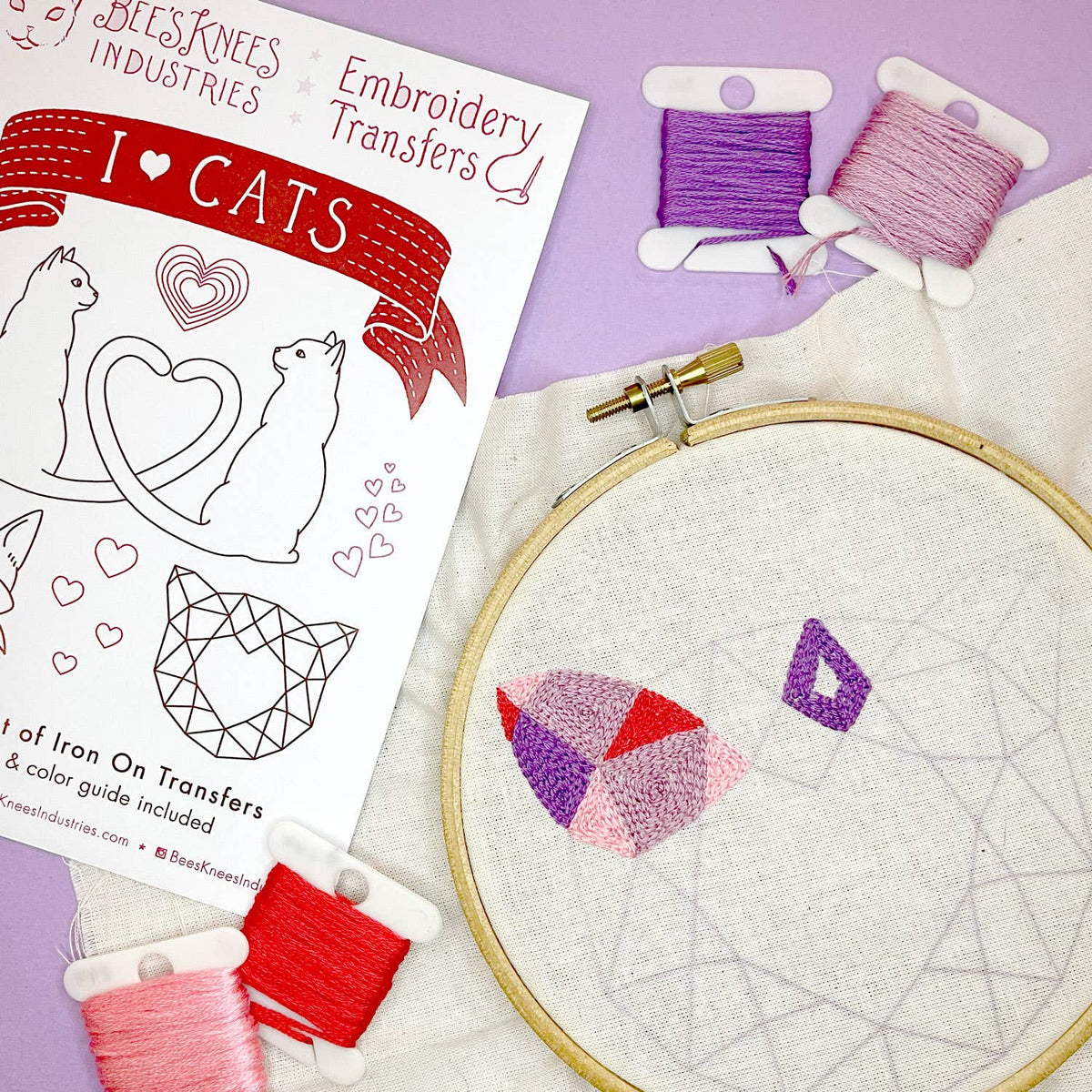Embroidery hoop with cat design, thread spools, and embroidery transfer paper on a purple background
