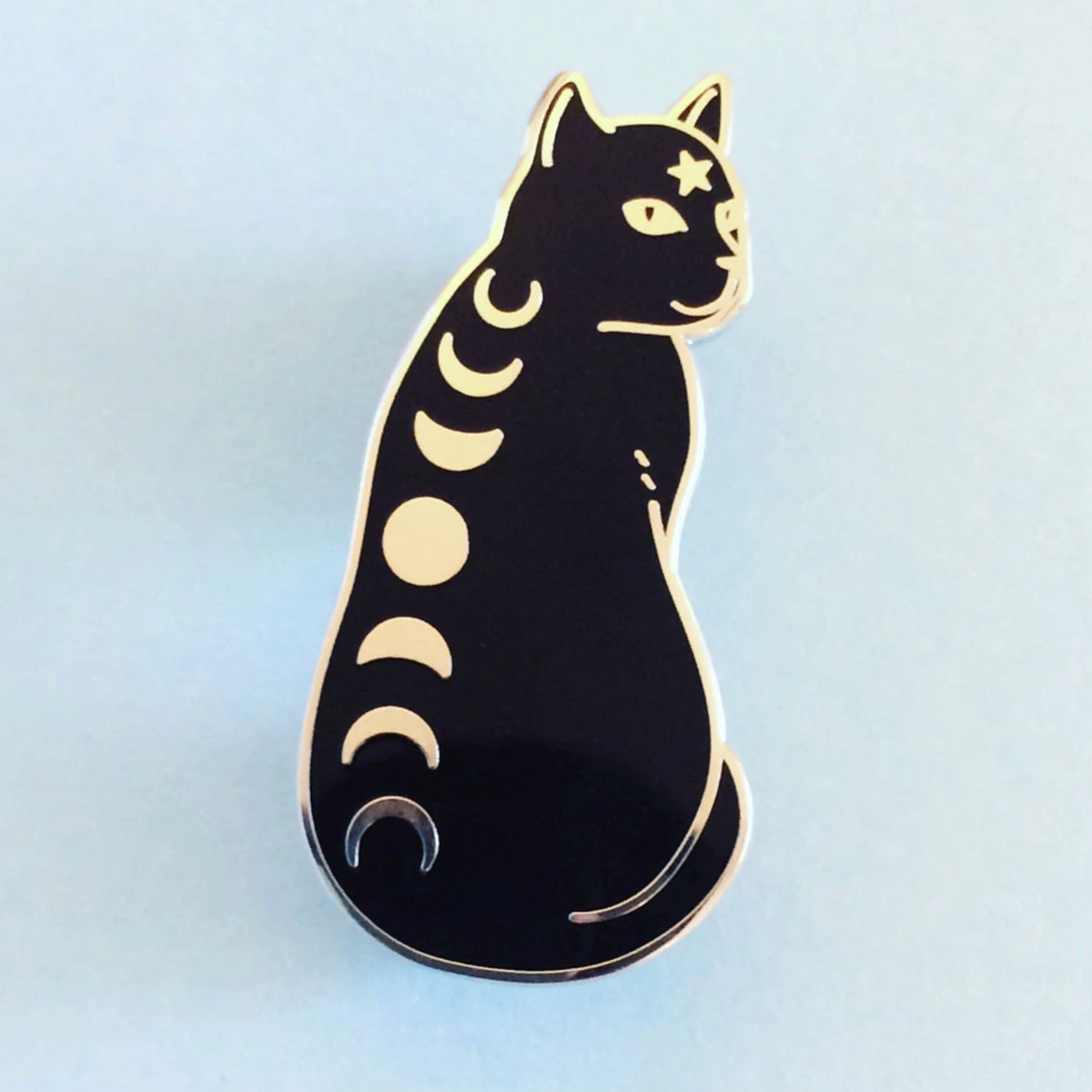 An enamel pin of a black cat with the phases of the moon down its back and a star on its forehead. 