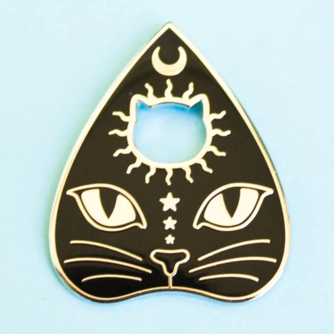 An enamel pin of a planchette with a cat's face on it and a crescent moon at the top. 