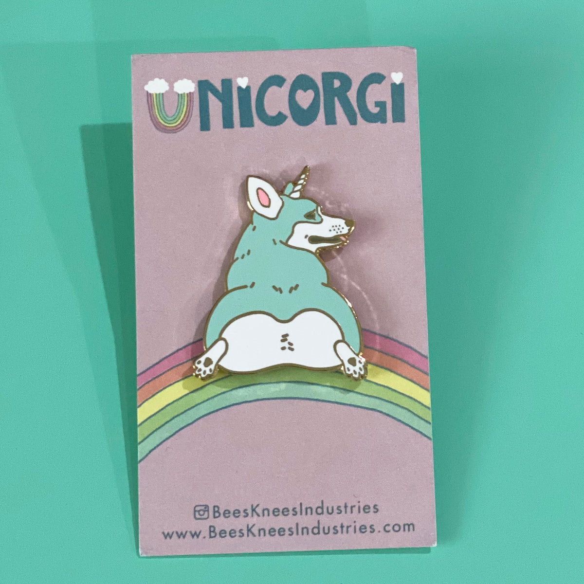An enamel pin of a mint colored corgi with a unicorn horn that has its butt facing the viewer on a backing card that says "UNICORGI" and has a rainbow. 
