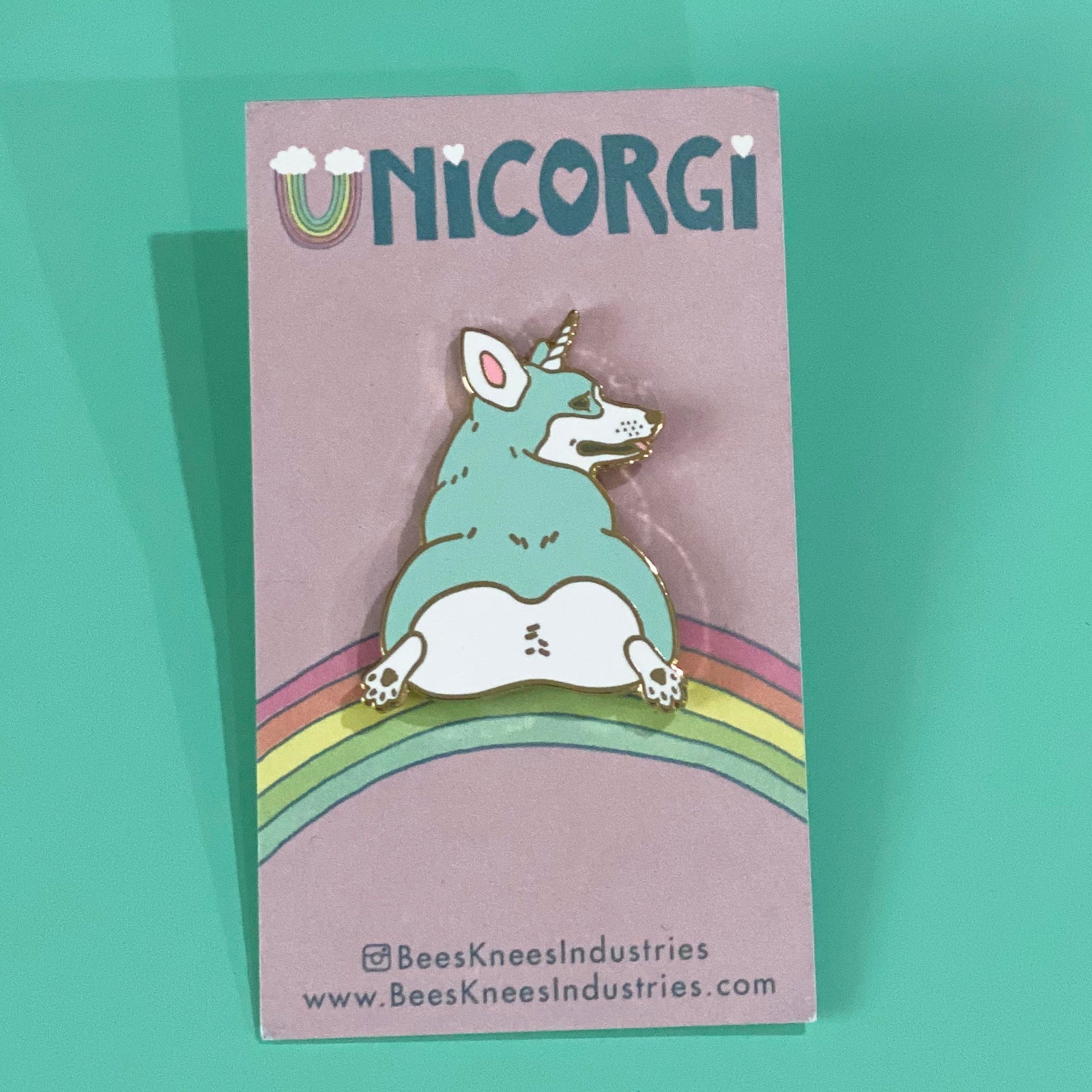 An enamel pin of a mint colored corgi with a unicorn horn that has its butt facing the viewer on a backing card that says "UNICORGI" and has a rainbow. 