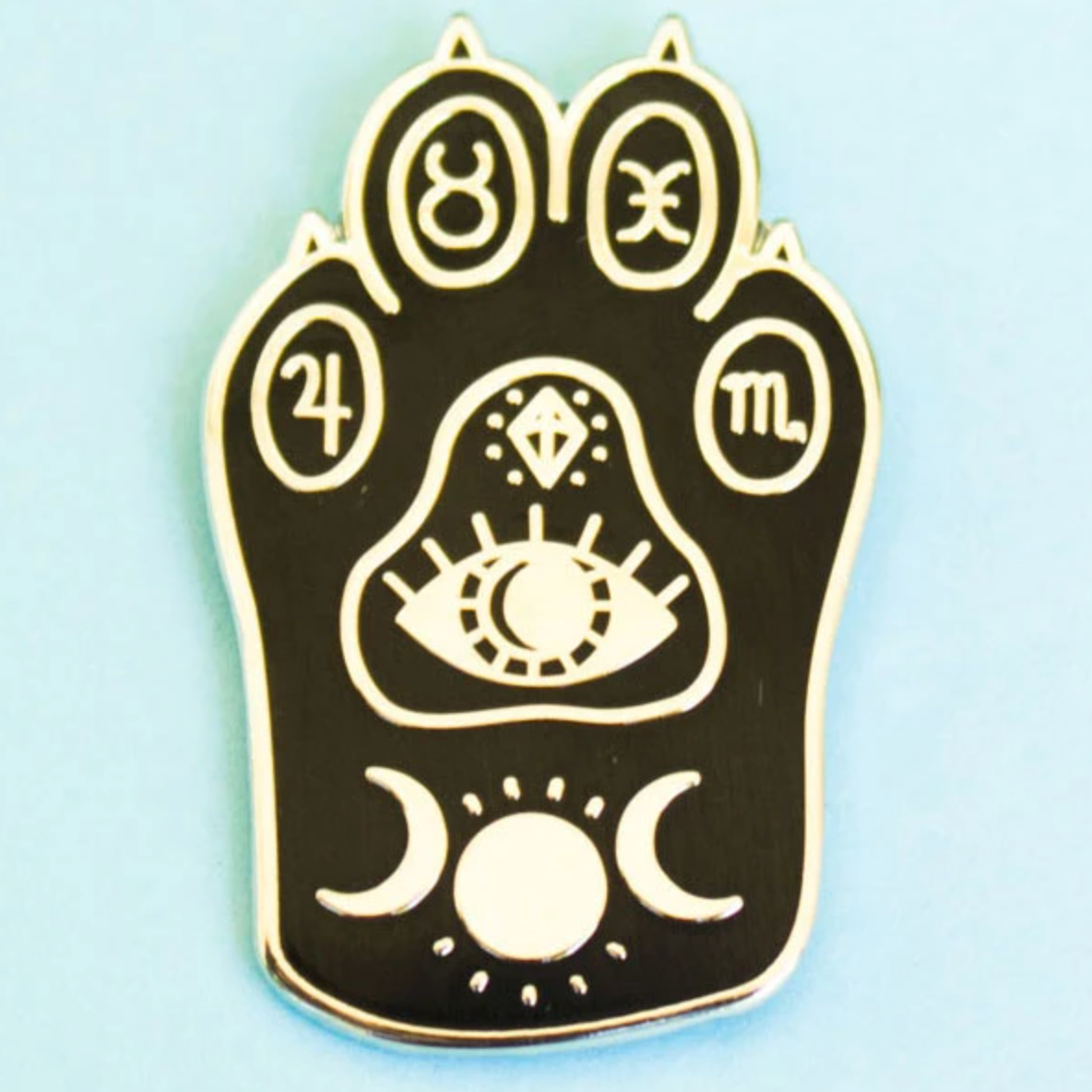 Enamel pin of a cat's paw with moon and sun designs, an eye, and zodiac signs on the toe pads. 