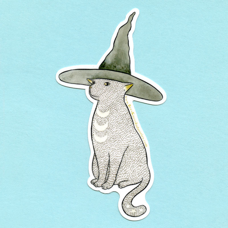 A sticker of a cat with three crescent moons on its chest, three stars on its tail, and a witch hat on its head. 