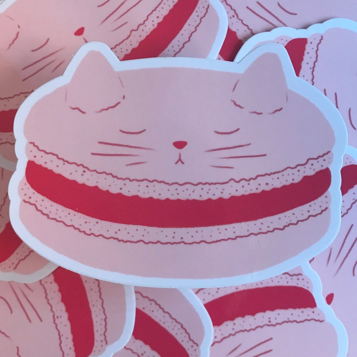 A sticker of a pink macaron with the face and ears of a cat on the top cookie. 
