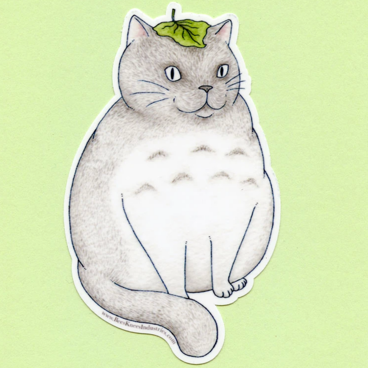 A sticker of a round gray cat with a leaf on its head against a green background.