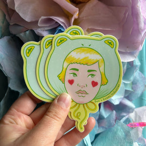 A hand holds a stack of die-cut stickers shaped like frog faces, featuring a stylized blonde woman's face with red heart-shaped cheeks. The stickers are shown against a background of soft purple and blue paper flowers.