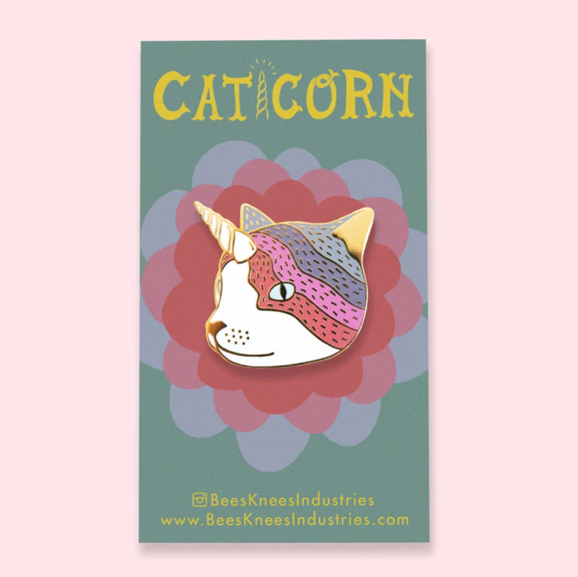 An enamel pin of a cat with a unicorn horn on a backing sheet that says "Caticorn" against a light pink background.
