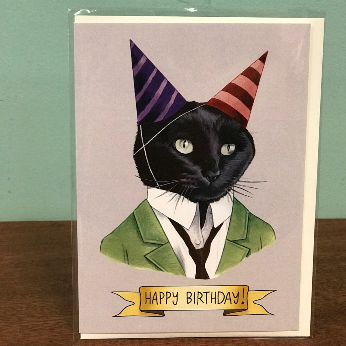 Birthday card with a black cat wearing a green suit and party hats on a beige background.