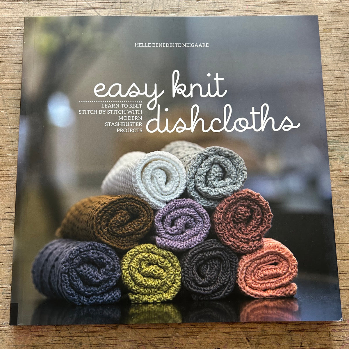 A book titled "easy knit dishcloths" by Helle Benedikte Negaard lies on a wooden surface. The cover shows a stack of knitted cloths in assorted colors, including cream, gray, brown, purple, yellow, black, and terracotta, arranged on a dark reflective surface.