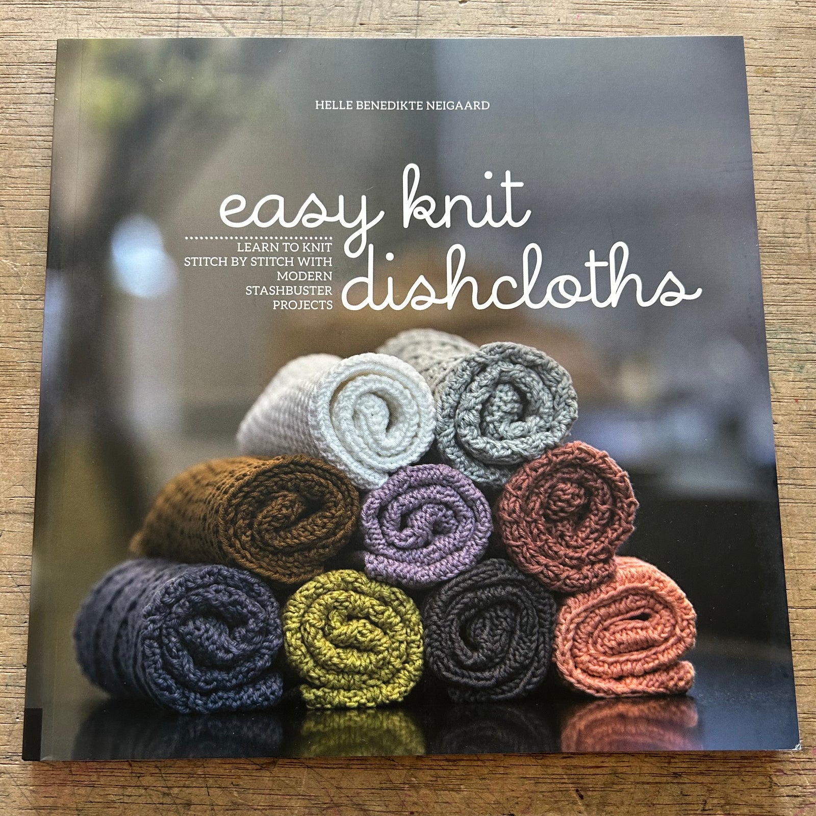 A book titled "easy knit dishcloths" by Helle Benedikte Negaard lies on a wooden surface. The cover shows a stack of knitted cloths in assorted colors, including cream, gray, brown, purple, yellow, black, and terracotta, arranged on a dark reflective surface.