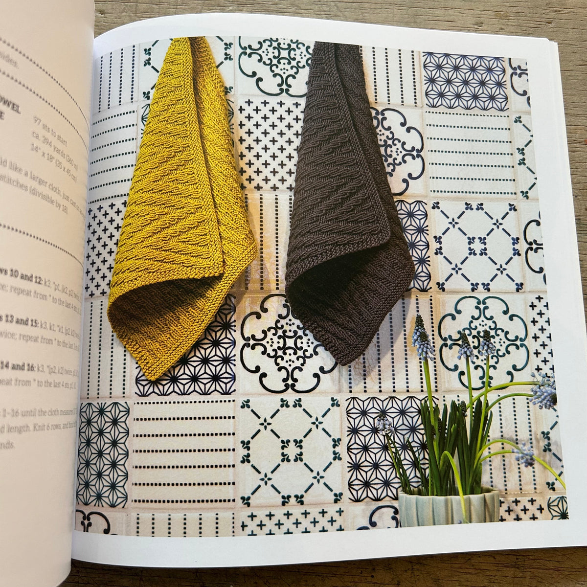 An open book showing a knit dishcloth in mustard yellow and another in dark brown, styled over a patchwork tile pattern background with a small potted plant.