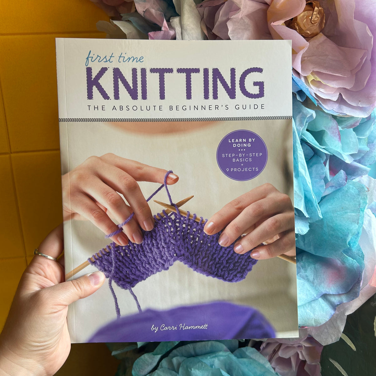 A person holds a book titled "first time KNITTING: THE ABSOLUTE BEGINNER'S GUIDE" with a purple knitted piece in progress shown on the cover. The book is photographed against a background of pastel flowers and a yellow surface.