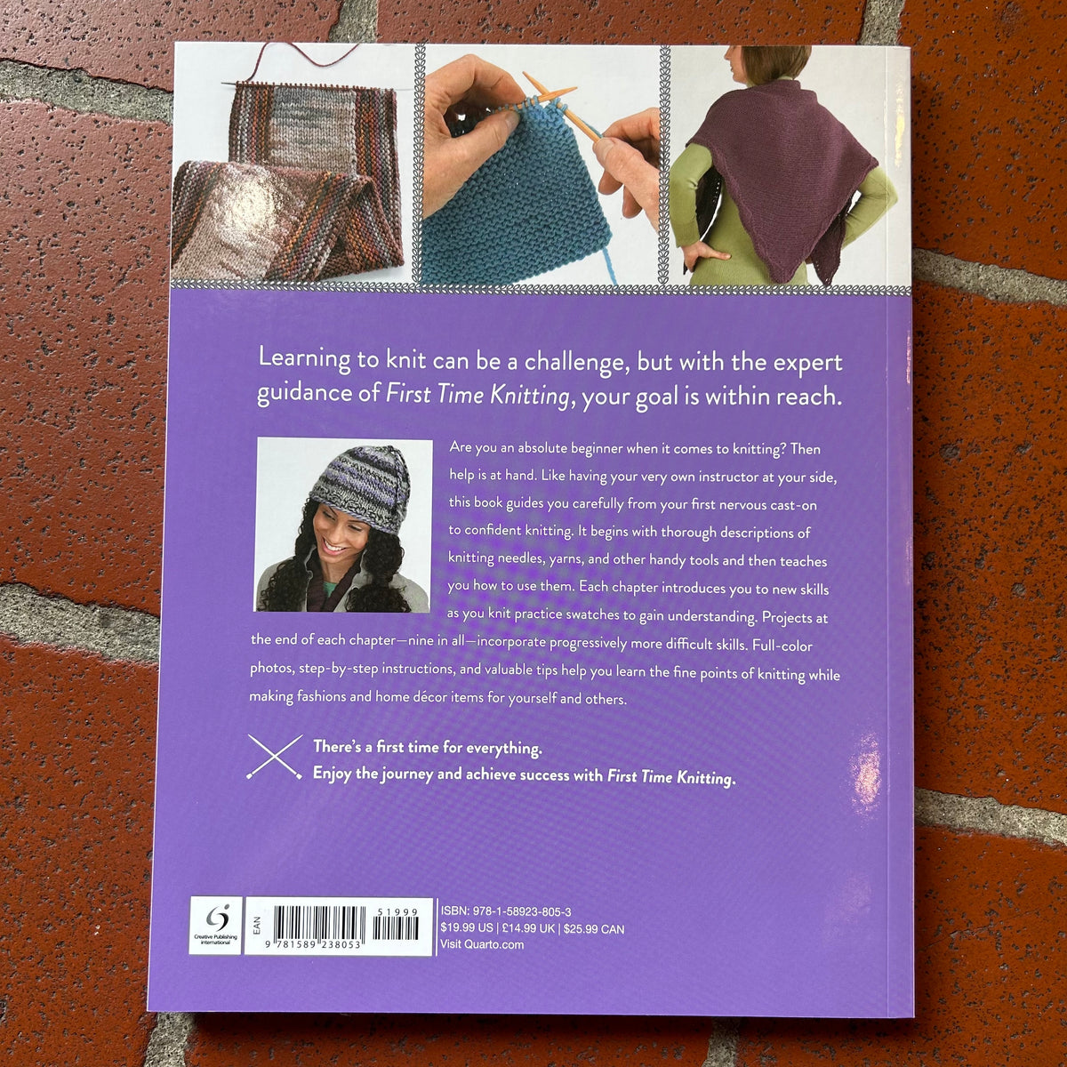 The back cover of the book 'First Time Knitting' featuring a purple background with a small photo of a knitted hat, a barcode, and a quote, displayed flat on a red brick surface.
