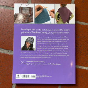 The back cover of the book 'First Time Knitting' featuring a purple background with a small photo of a knitted hat, a barcode, and a quote, displayed flat on a red brick surface.