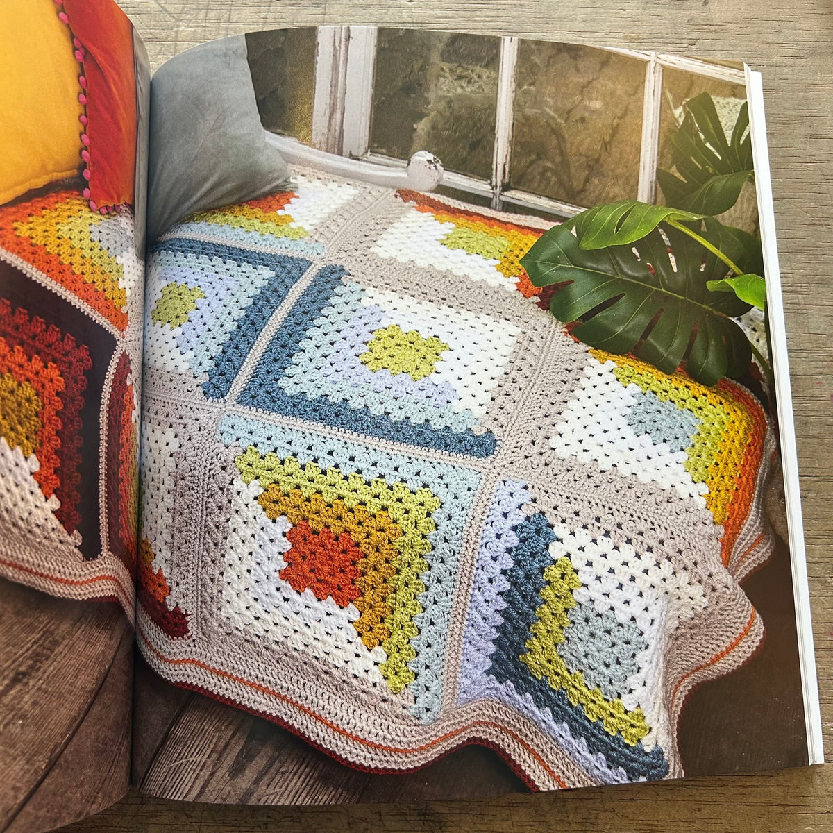 An open book showing a crocheted granny-square throw blanket draped over a bed. The blanket features multiple colorful squares in cream, blue, yellow, orange, and gray.