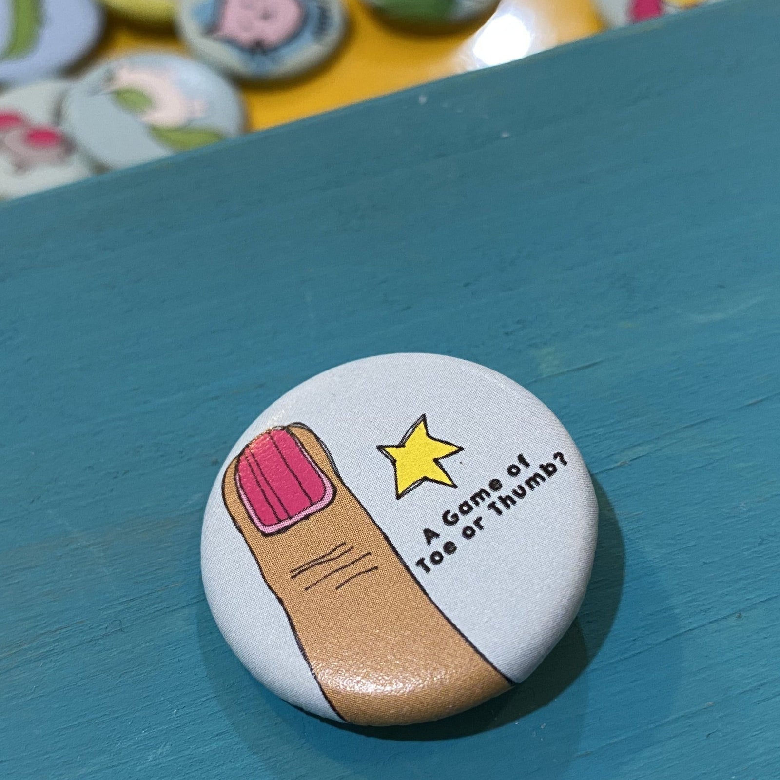 Button badge with a cartoon hand and text  that says "A Game of Toe or Thumb?"
 on a blue surface