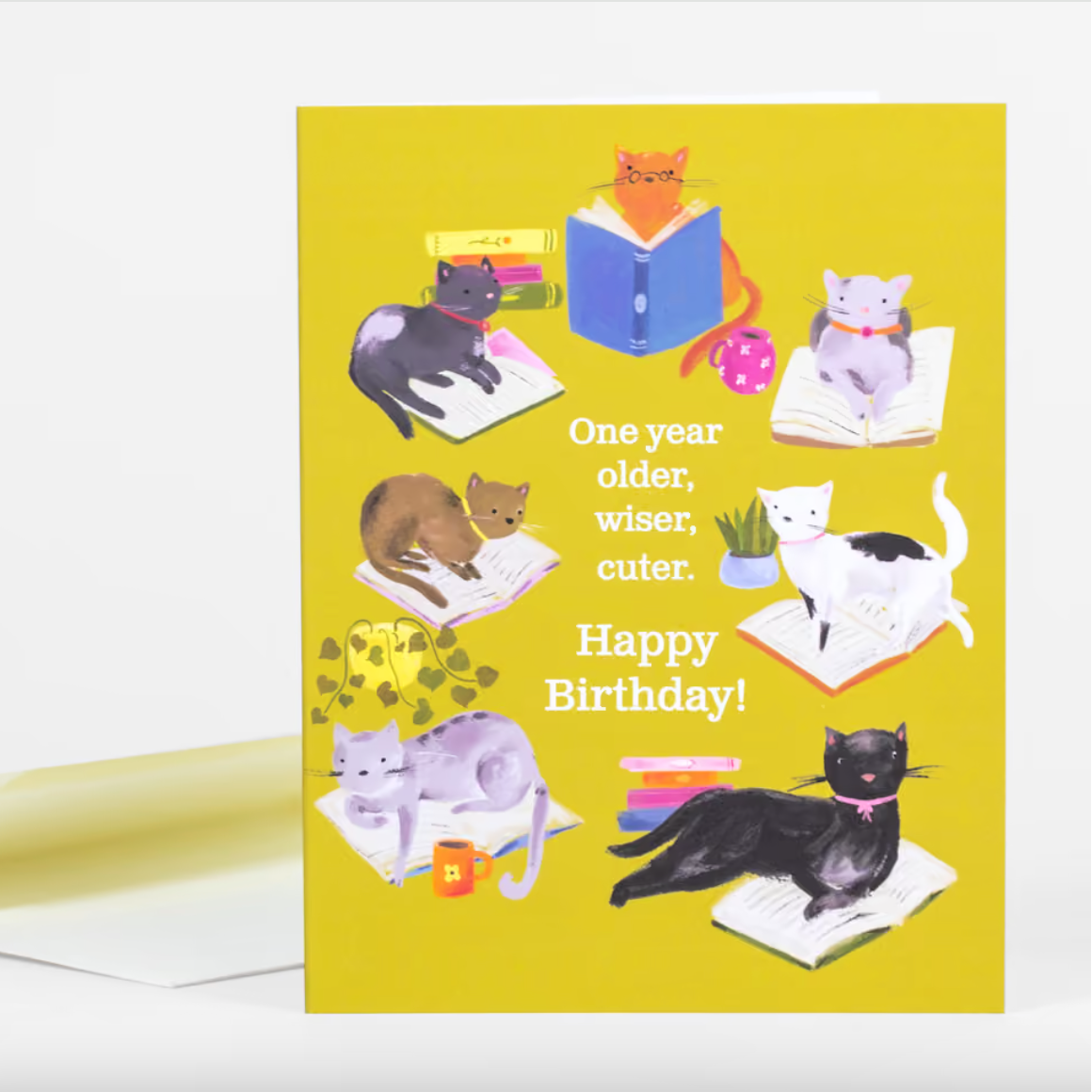 A yellow card with a group of cats lounging on books with some houseplants and text that says "One year older, wiser, cuter. Happy Birthday!"