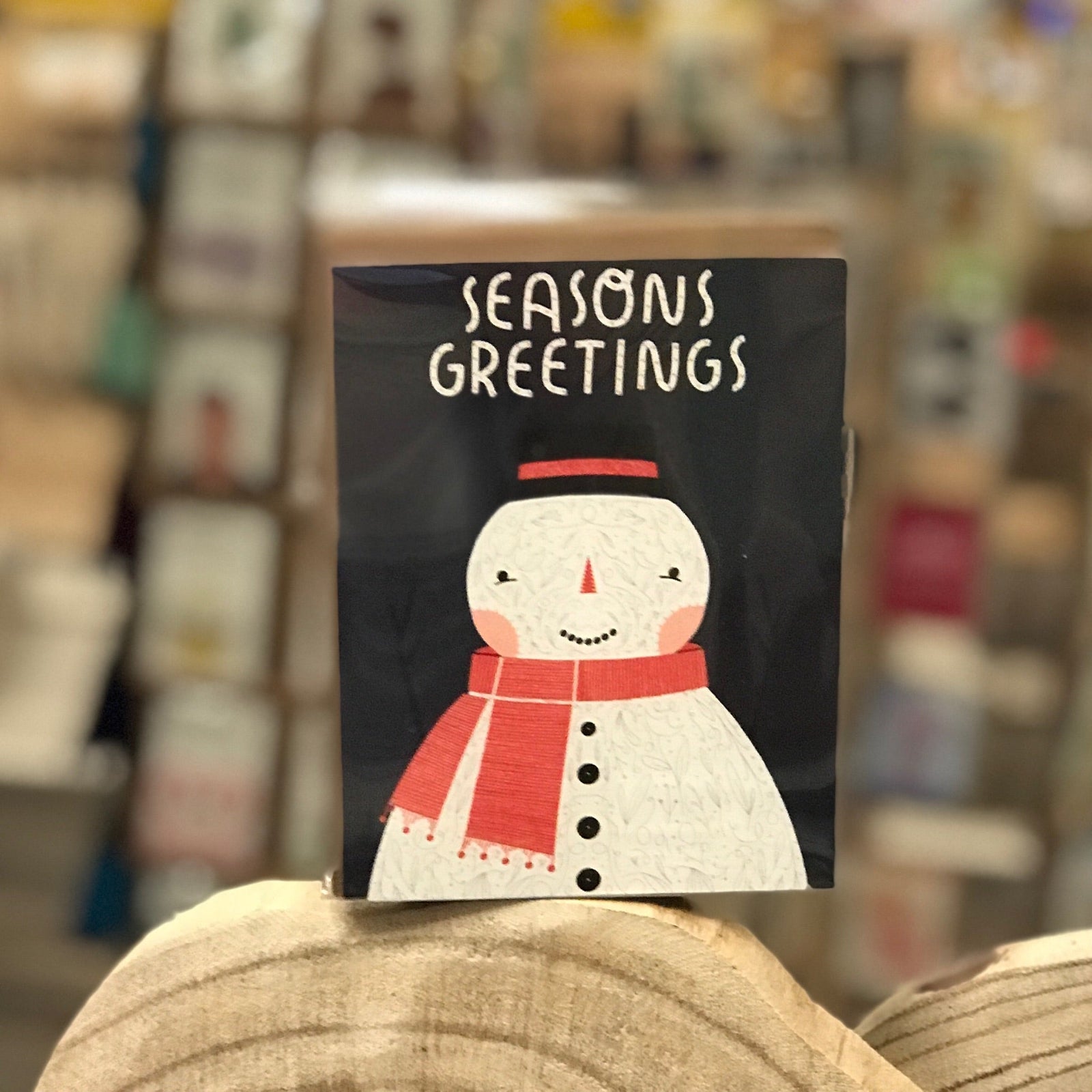 Holiday greeting card with a snowman wearing a top hat and a red scarf. The card has a black background and is shown. in a retail setting. 