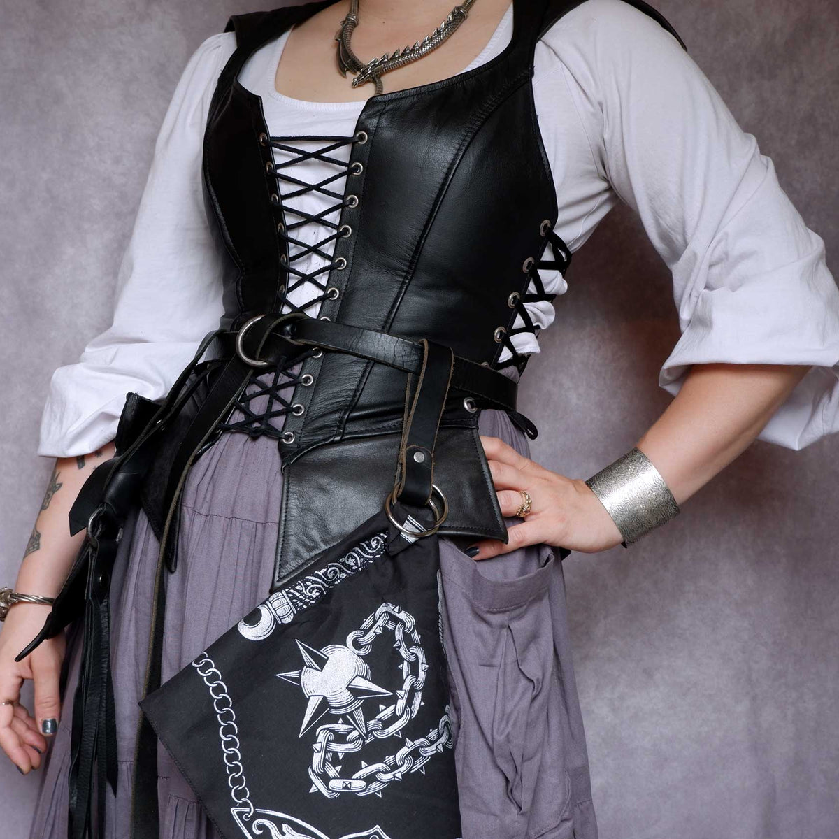 Person wearing a black leather corset that has a black bandana with silver designs tied to it against a gray background