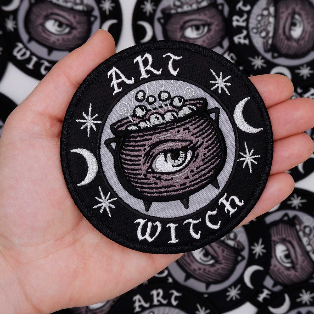 Hand holding a 'Art Witch' patch with a cauldron and eye design against a patterned background.