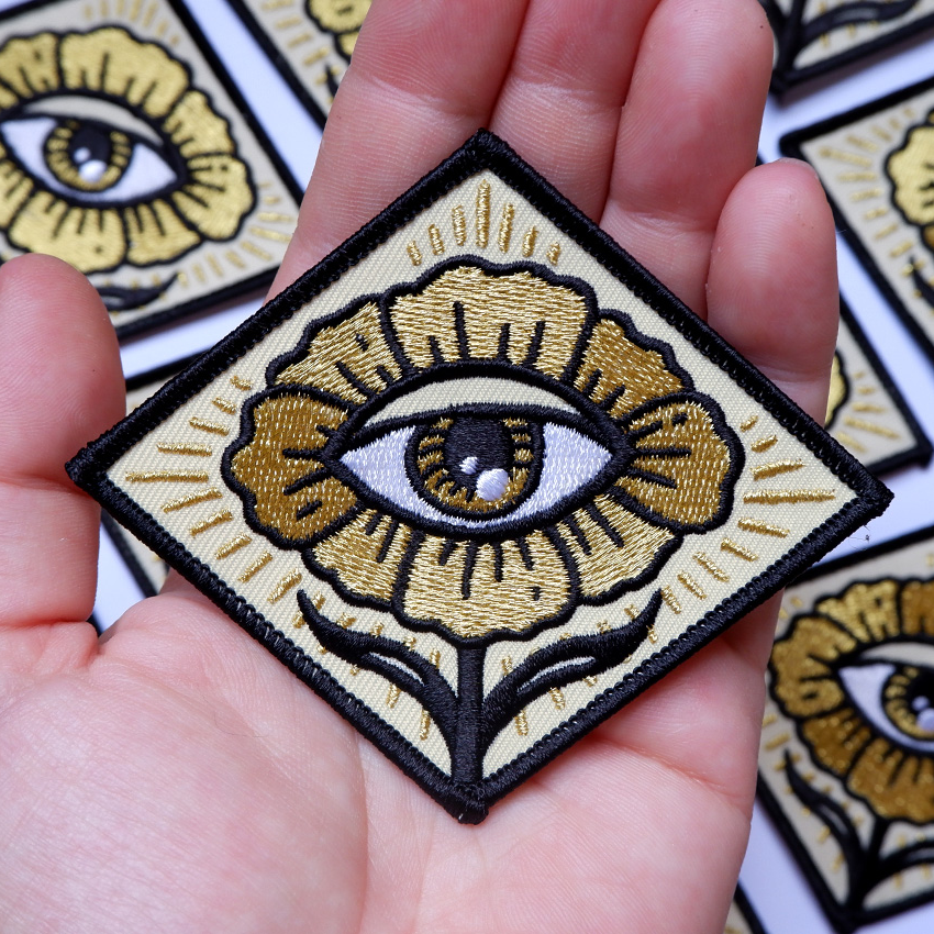 A gold diamond-shaped patch with a design of flower with an eye in the center. 