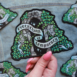 Decorative patch with 'Everything is temporary' text held by a hand on a textured surface.