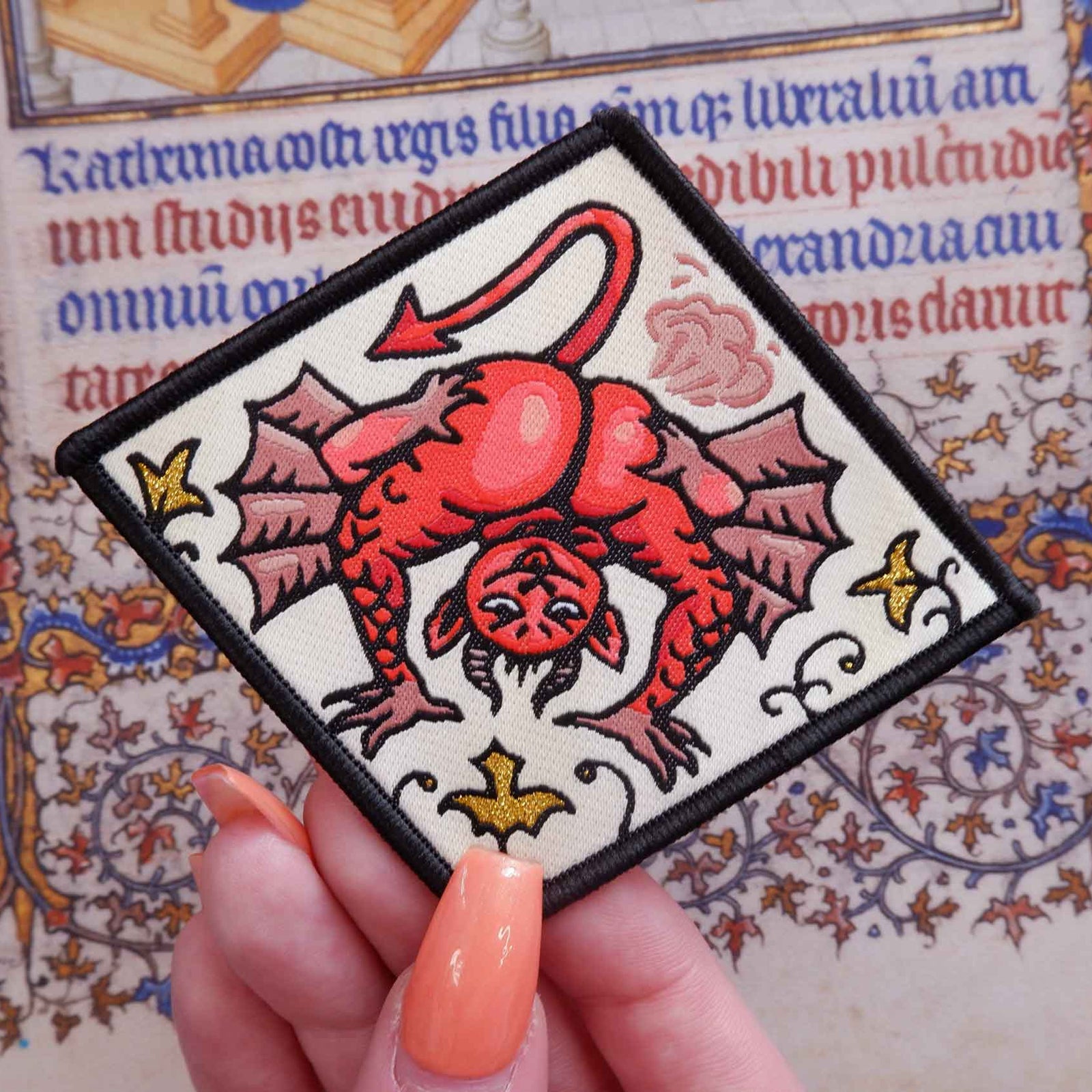 A hand with peach-colored nails holds a square embroidered patch featuring a red devil illustration with pink wings and gold stars, set against a background of a vintage-style illuminated manuscript.