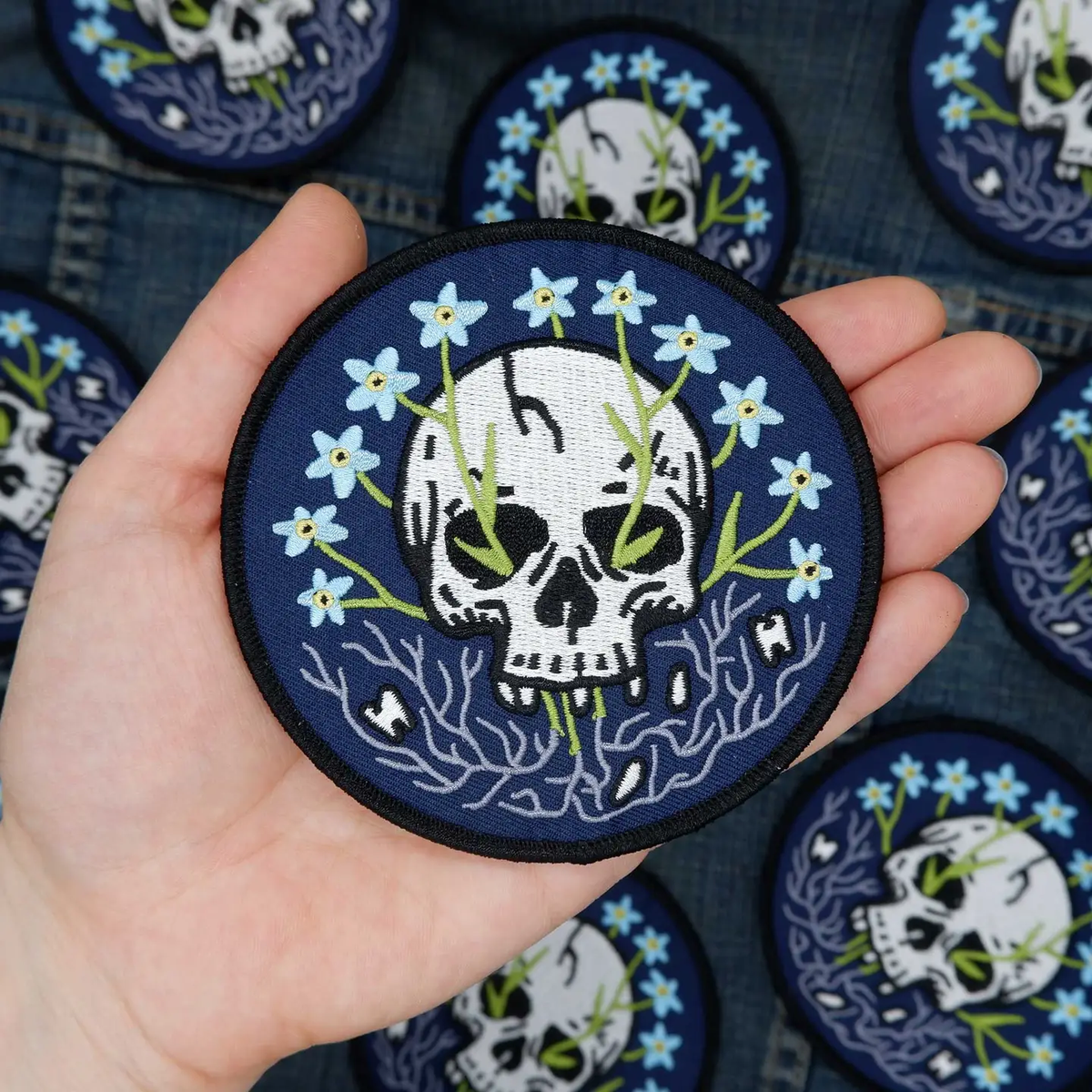 A round blue patch featuring a skull with forget-me-not flowers growing out of it. 