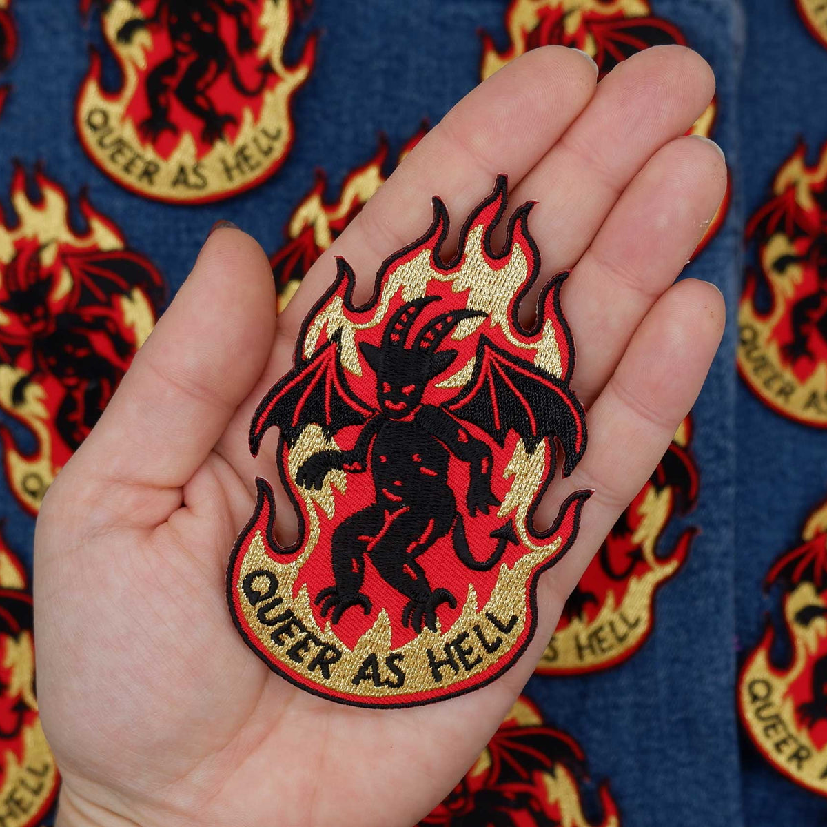 Hand holding a patch with a devil design and 'Queer as Hell' text against a patterned background.