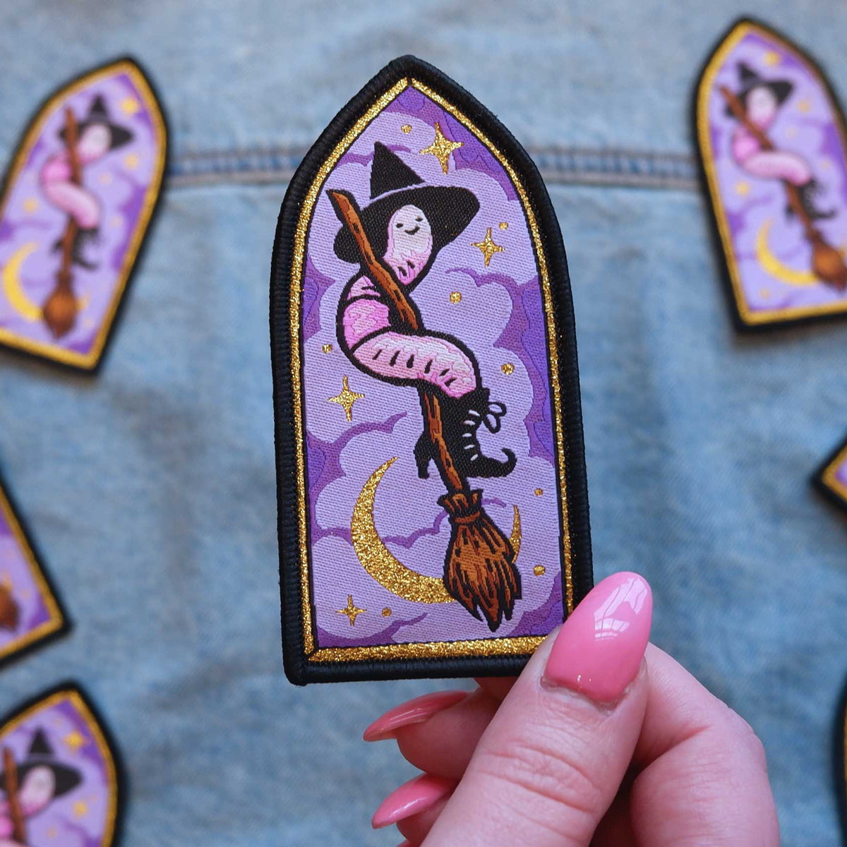 Patch of a worm witch on a broomstick held by a hand with pink nail polish, surrounded by similar patches on a textured background.