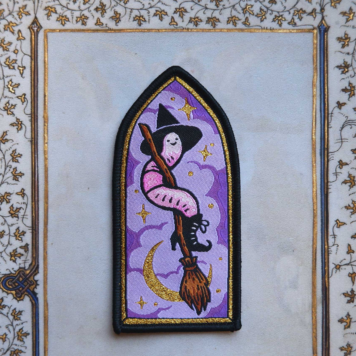 Embroidered patch of a worm witch on a broomstick with a purple background on a decorative frame.