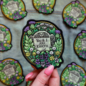 Decorative patch with 'You & I Are Earth' text held by a hand on a patterned background.