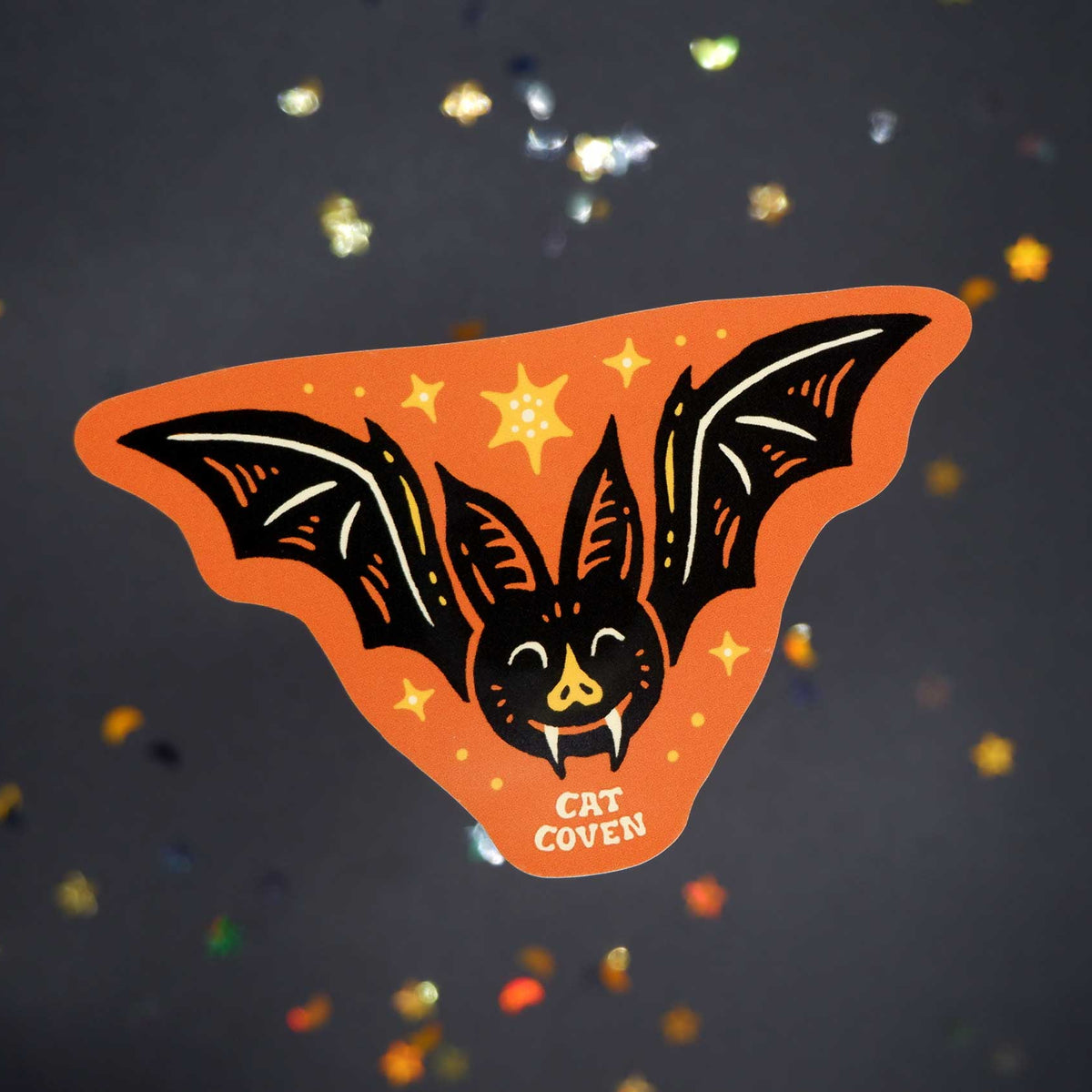 An orange bat-shaped sticker with a black bat illustration, small yellow stars, and the words 'CAT COVEN' at the bottom, shown on a dark glittery background.