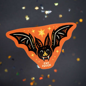 An orange bat-shaped sticker with a black bat illustration, small yellow stars, and the words 'CAT COVEN' at the bottom, shown on a dark glittery background.