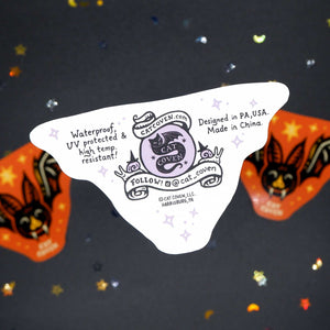 A white die-cut sticker featuring a purple cat logo and text, displayed on a dark glittery background with orange bat-shaped sticker.